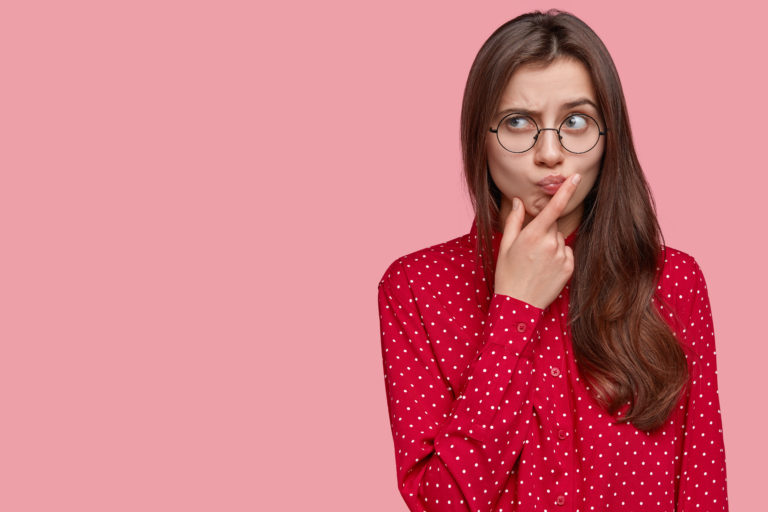 Young attractive woman with thoughtful expression, thinks about question, feels doubt, raises eyebrows, holds chin, dressed in formal clothes, isolated over pink background with free space on left artículo 28