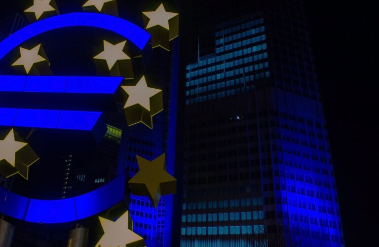 Escultura iluminada del símbolo del euro con estrellas amarillas sobre un fondo de edificios modernos por la noche, resaltando la importancia de los depósitos financieros en la economía europea.