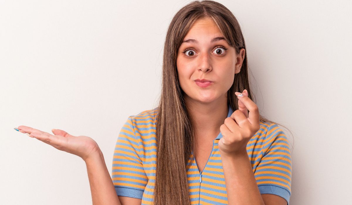 Una mujer joven, de pelo largo, encogiéndose de hombros y gesticulando con los dedos, está de pie sobre un fondo blanquecino, contemplando su plan de pensiones.