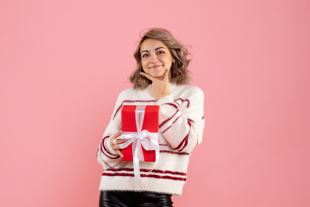 Una mujer sonriente, llena de alegría y expectativa, sostiene una caja de regalo roja adornada con un lazo blanco sobre un fondo rosa. Es como si acabara de recibir la sorpresa perfecta por sus esfuerzos reflejados en la nómina de este mes.