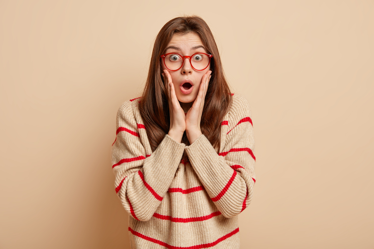 Una persona con gafas rojas y un jersey de rayas parece sorprendida, con las manos en las mejillas, sobre un fondo beige. El intrincado patrón recuerda a letras en espiral, lo que añade un giro inesperado a la escena.