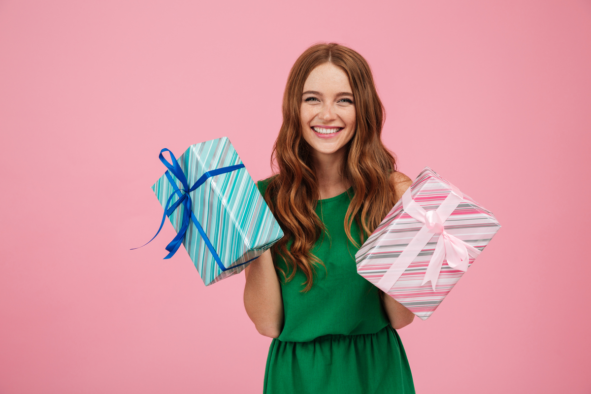 Una mujer sonriente con un vestido verde, cuya alegría es tan brillante como el fondo rosa, sostiene dos cajas de regalo, tal vez celebrando su última nómina con un toque de indulgencia.