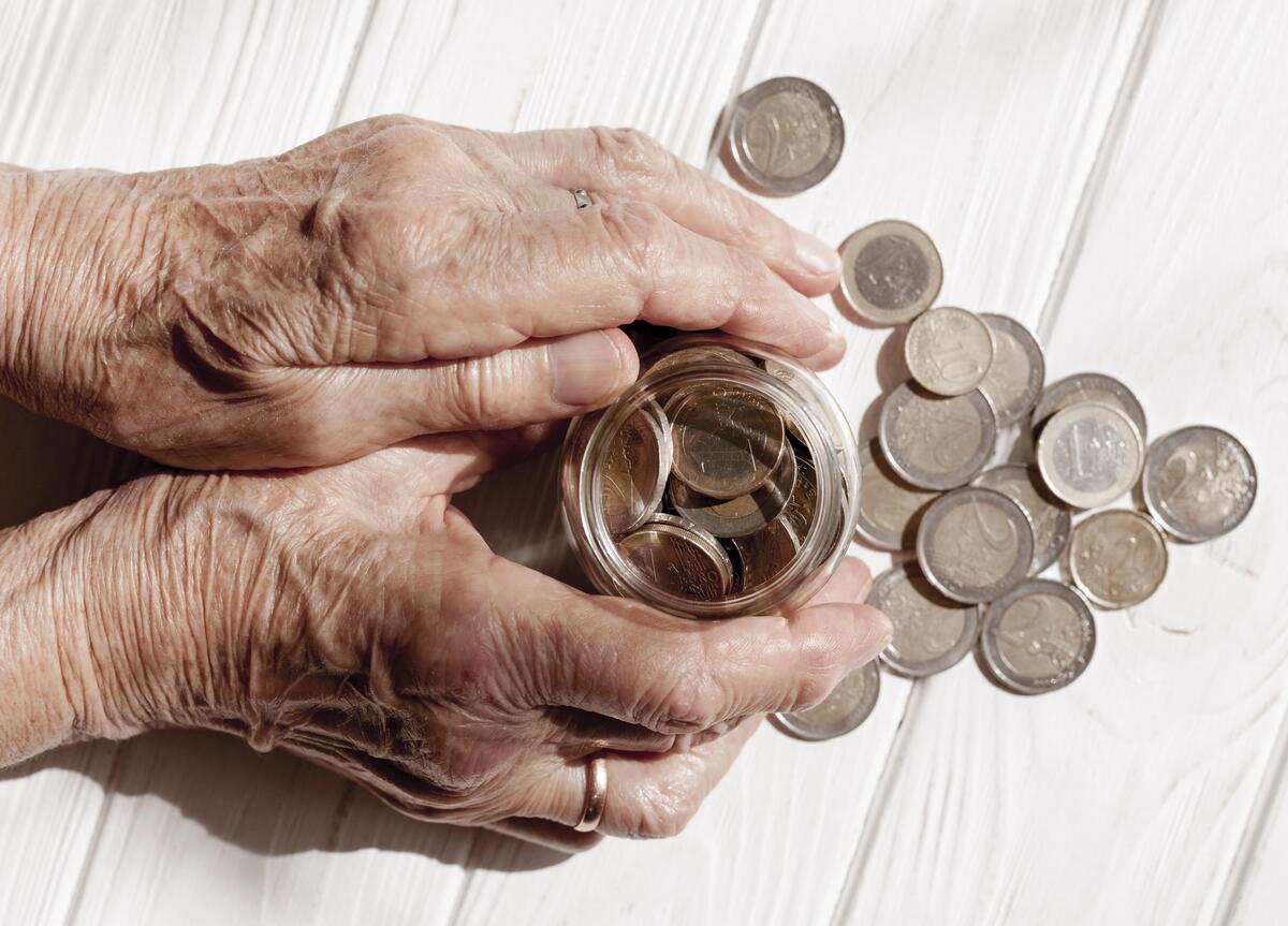 Unas manos ancianas sostienen con delicadeza un frasco de monedas, con más monedas esparcidas sobre una superficie de madera blanca, simbolizando los cuidadosos ahorros vinculados a planes de pensiones vitales.