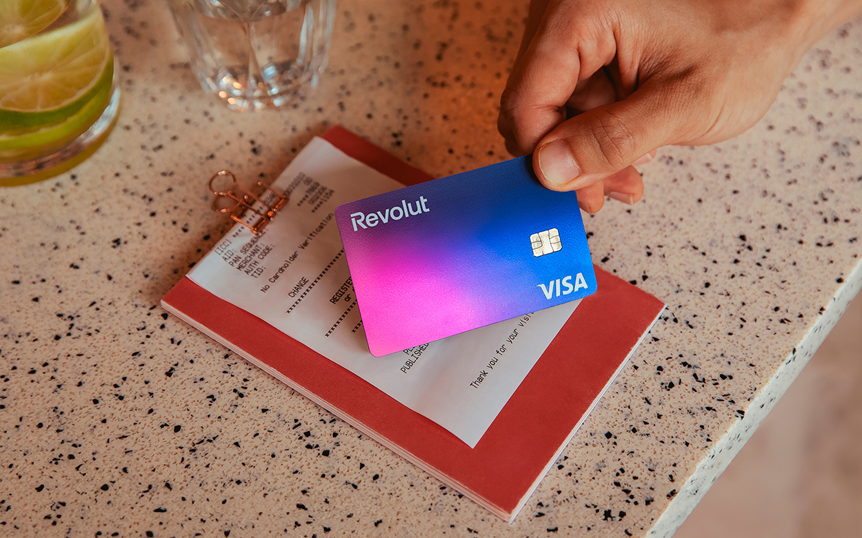 Una persona sostiene orgullosamente una colorida tarjeta Visa de Revolut sobre un portapapeles rojo en un mostrador moteado, ilustrando cómo el plan de recompensas realmente vale la pena.