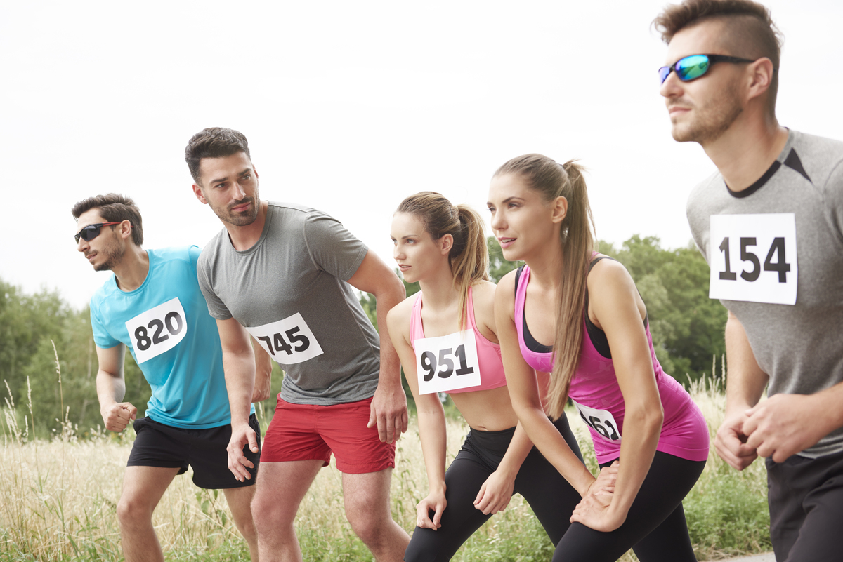 Cinco corredores con dorsales numerados se preparan para iniciar una carrera al aire libre en un campo de césped, mientras sus métricas de rendimiento son monitoreadas por un avanzado sistema roboadvisor.