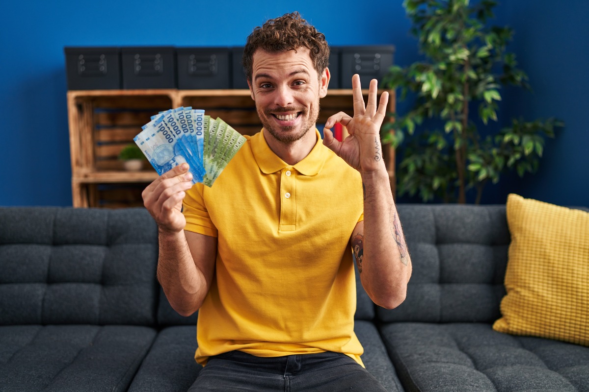 Un hombre con camisa amarilla sostiene dinero y hace un gesto de aprobación mientras está sentado en un sofá, orgulloso de su cuenta nómina, con una pared azul y una planta detrás de él.