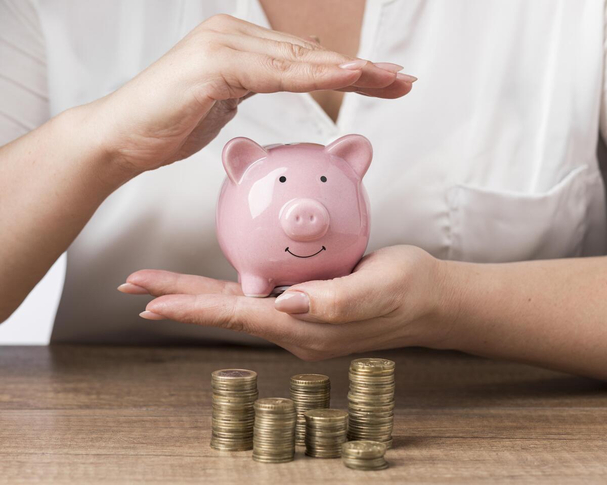 Una persona que sostiene una alcancía rosa, rodeada de pilas de monedas sobre una mesa de madera, reflexiona sobre el mejor momento para empezar a ahorrar para la jubilación.
