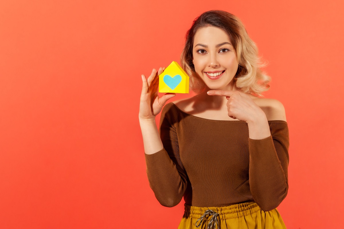 Una mujer sonriente, que representa la esperanza de los jóvenes, sostiene una pequeña maqueta de casa con un corazón azul sobre un fondo naranja, que simboliza el sueño de opciones accesibles de financiación e hipoteca para compradores de vivienda por primera vez.
