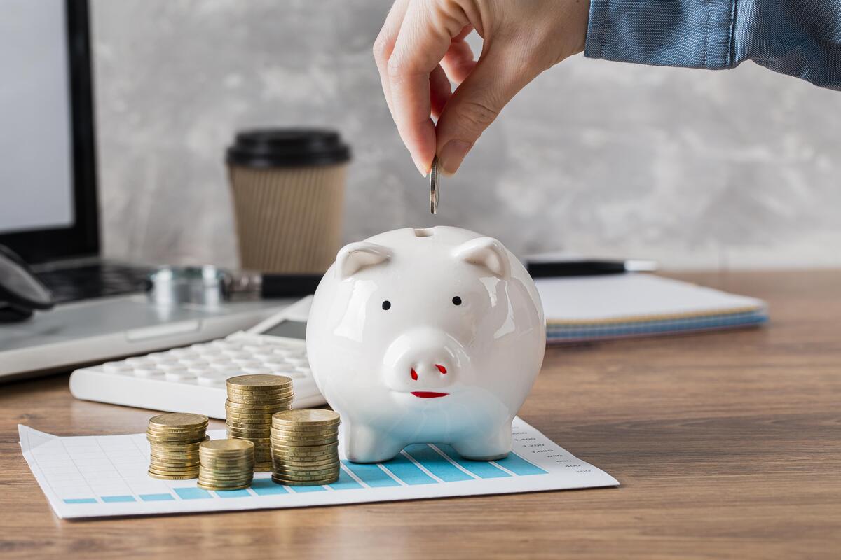 Una mano deposita una moneda en una alcancía sobre un escritorio adornado con monedas apiladas y artículos de oficina esenciales, simbolizando los beneficios de una cuenta remunerada.