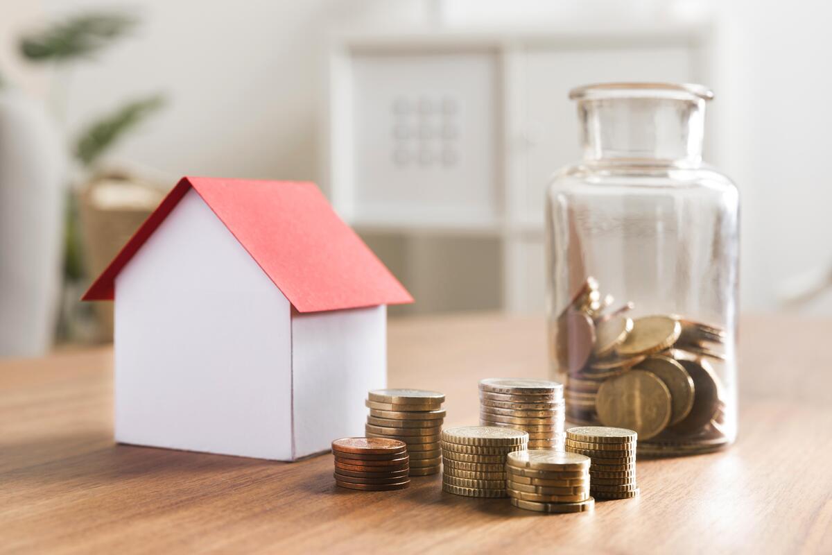 Una pequeña maqueta de casa, monedas apiladas y un tarro de monedas descansan sobre una mesa de madera, ilustrando la diferencia a la hora de ahorrar para una vivienda o considerar hipotecas.