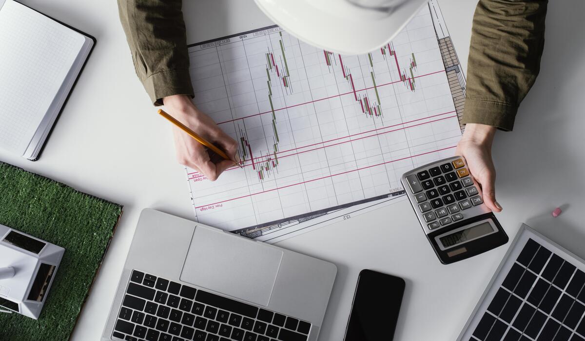Persona que analiza gráficos en papel, usando una calculadora y un lápiz, con una computadora portátil y un teléfono en el escritorio, administrando de manera eficiente los datos de PIAS para obtener información precisa.