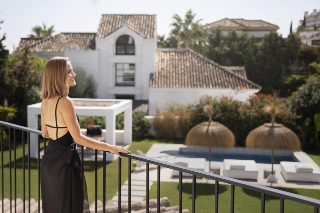 Una mujer vestida de negro se encuentra en un balcón, con vistas a un jardín con piscina y sombrillas de paja, tal vez soñando con su próxima casa o planificando su futura hipoteca.