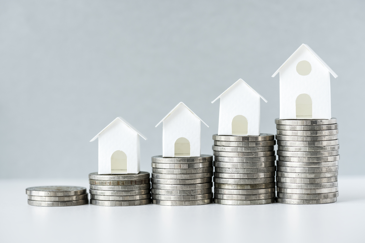 Monedas apiladas con alturas crecientes, cada una rematada con una pequeña vivienda blanca, que simboliza el aumento del valor de la propiedad.