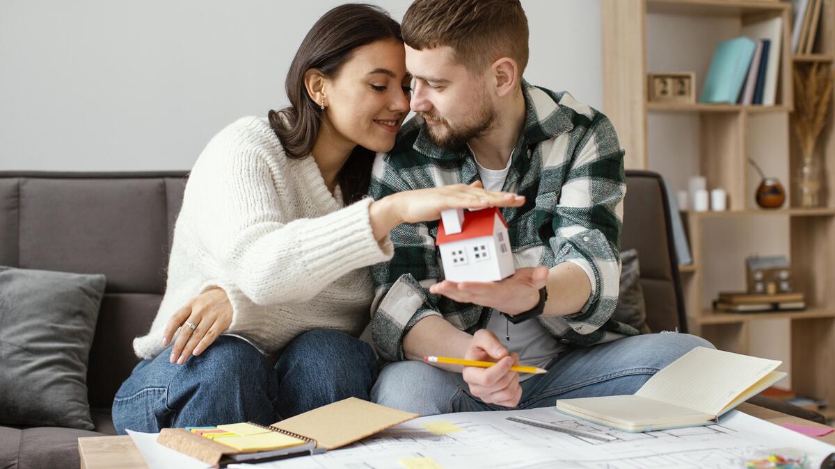 Una pareja se sienta en un sofá, sonriendo y sosteniendo una maqueta de casa sobre planos y cuadernos, planificando la casa de sus sueños con la ayuda de Aval.