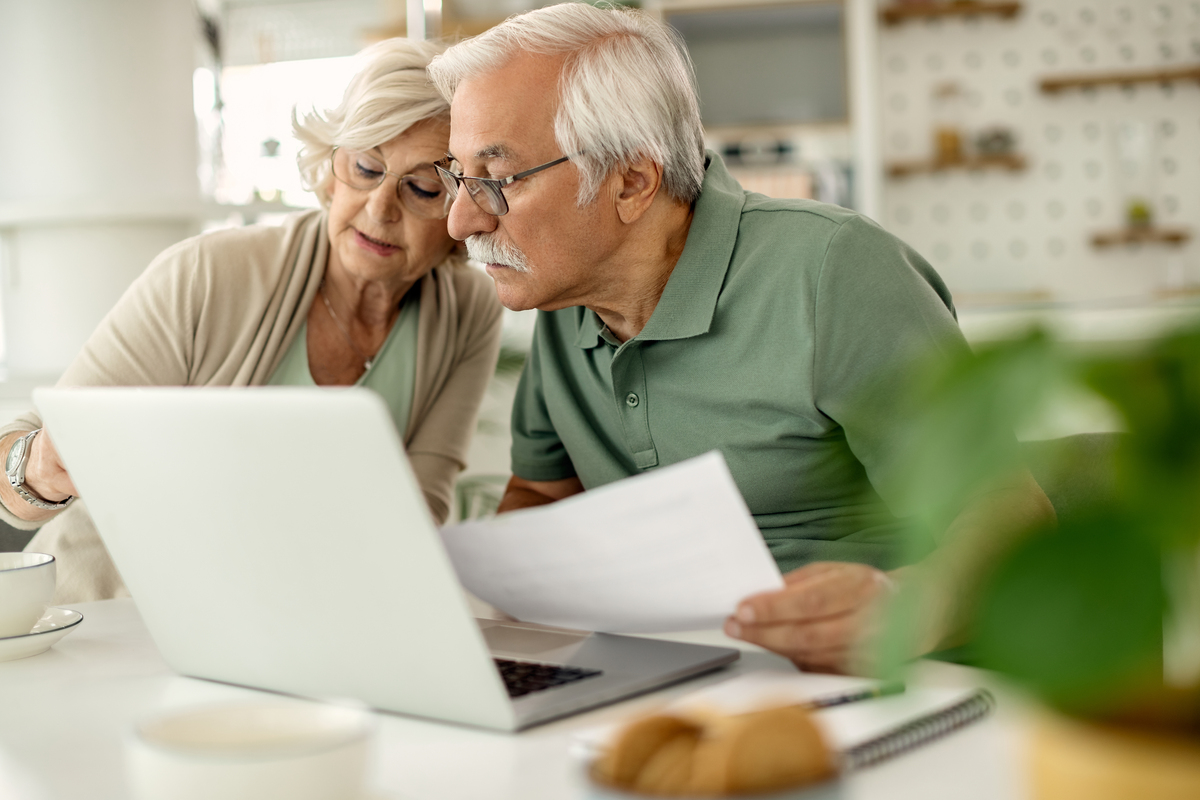 Pareja mayor sentada en una mesa, mirando una computadora portátil y papeles juntos mientras revisan sus planes de pensiones.
