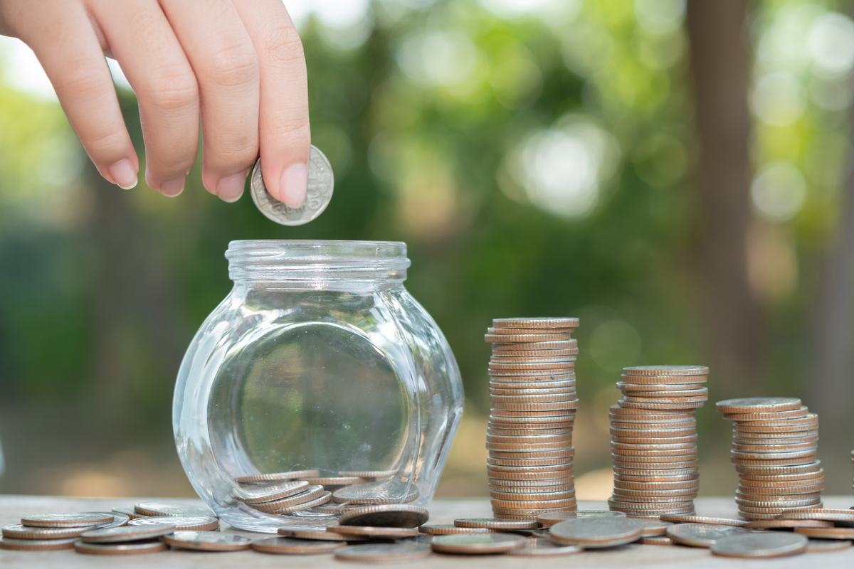 Mano colocando una moneda en un frasco de vidrio con monedas apiladas al lado en una mesa al aire libre, lo que representa el ahorro con los mejores depósitos.