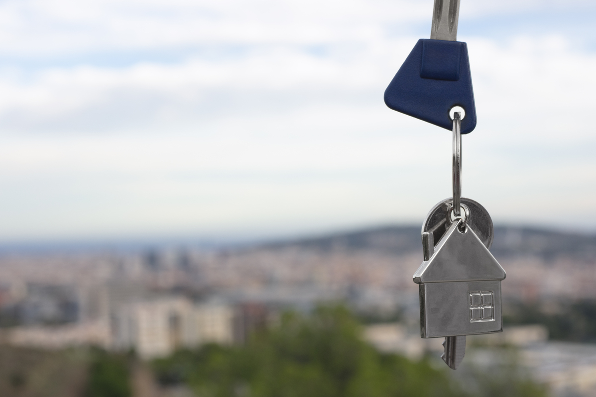 Un llavero con forma de casa y llaves colgando con un paisaje urbano difuminado al fondo, reflejando debates sobre pisos turísticos y cómo las ciudades buscan aumentar impuestos para gestionar viviendas vacías.