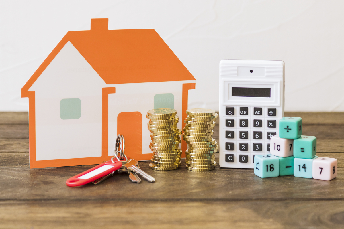 Casa de papel, llaves, monedas apiladas, calculadora y dados numerados sobre una mesa de madera, simbolizando la vivienda y las finanzas del hogar.