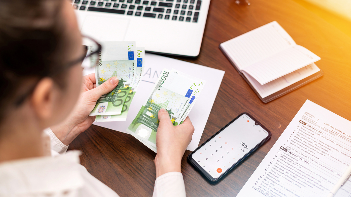 Persona contando billetes de euro en un escritorio con un ordenador portátil, una calculadora de teléfono, documentos y un cuaderno mientras revisa su nómina.