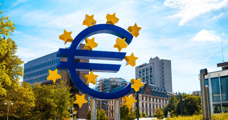 Gran símbolo del euro con estrellas amarillas delante de edificios de la ciudad, árboles y un cielo azul con nubes, que representa el impacto de las políticas del BCE y las tasas del euríbor en Europa.