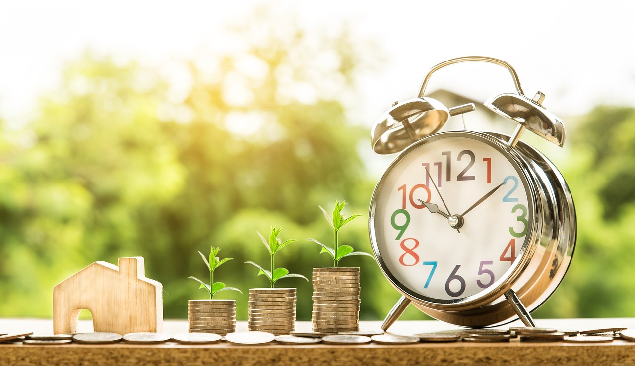 Reloj despertador, monedas apiladas con plantas y una pequeña maqueta de casa sobre una mesa al aire libre, simbolizando la rentabilidad y el crecimiento que puede derivar de un buen depósito a lo largo del tiempo.