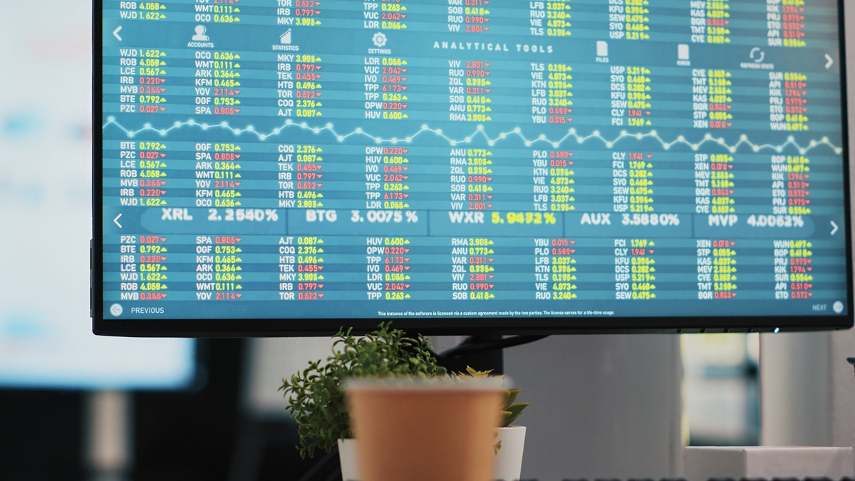 Un monitor de computadora muestra datos financieros y gráficos coloridos, mostrando un tablero de control de un roboadvisor; una planta y una taza de café están en primer plano.