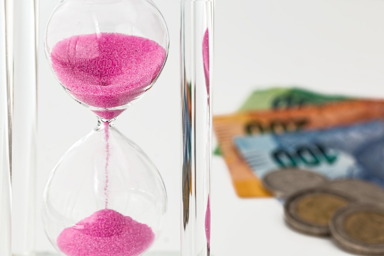 Reloj de arena con arena rosa junto a billetes y monedas de euro sobre una superficie blanca, simbolizando la importancia del tiempo en la gestión de los depósitos.