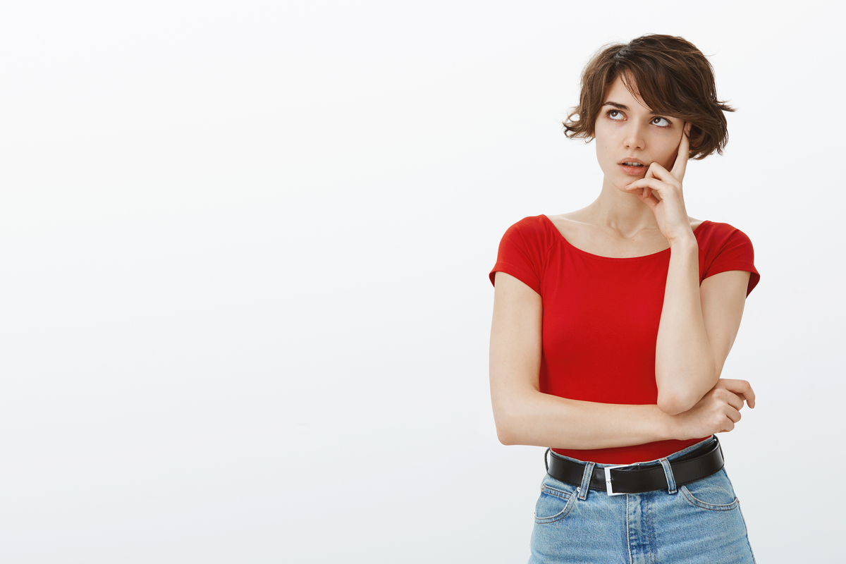 Mujer joven con una camisa roja y pantalones vaqueros de pie, con mirada pensativa, con la mano en la barbilla contra un fondo blanco, tal vez contemplando oportunidades de vivienda jóvenes.