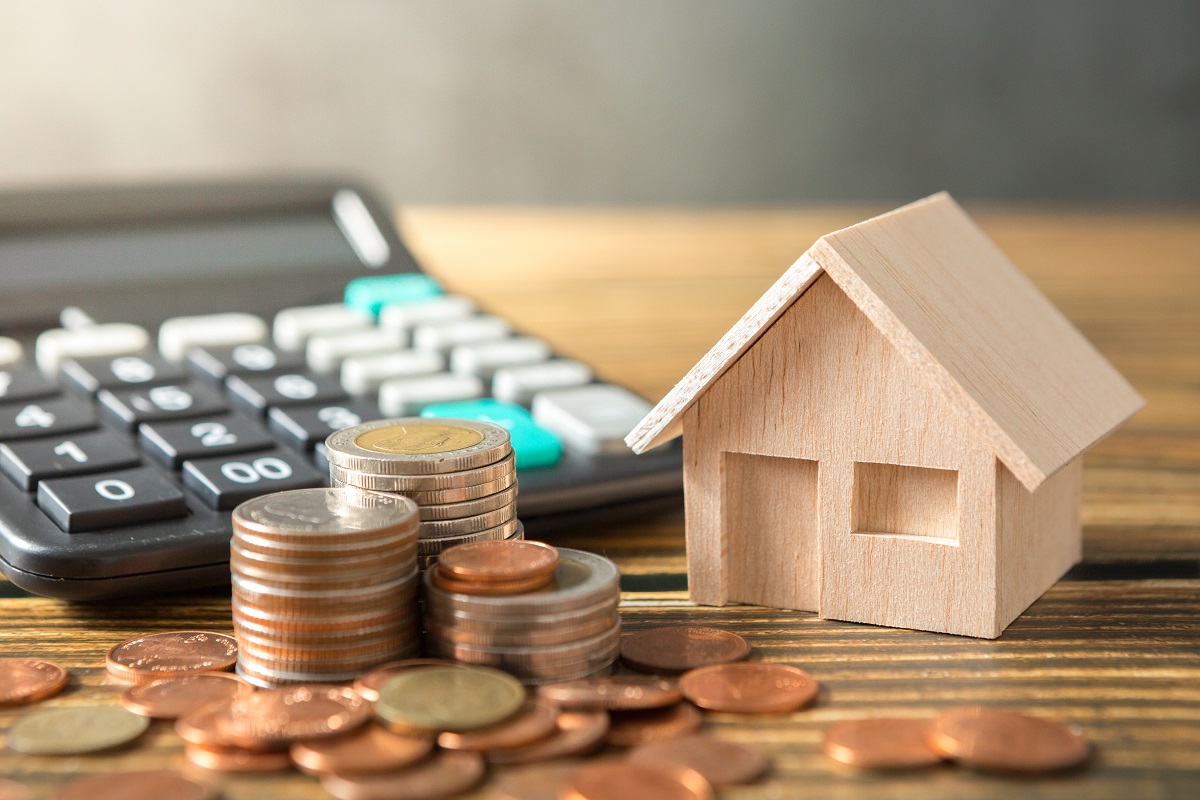 Pequeña casa de madera, monedas y una calculadora sobre una mesa de madera, simbolizando las finanzas o el presupuesto doméstico en junio mientras los tipos del euríbor fluctúan.