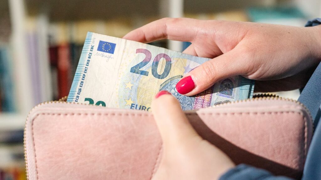 Una mano con uñas rosas introduce billetes de euro, o dinero en efectivo, en una cartera rosa claro.