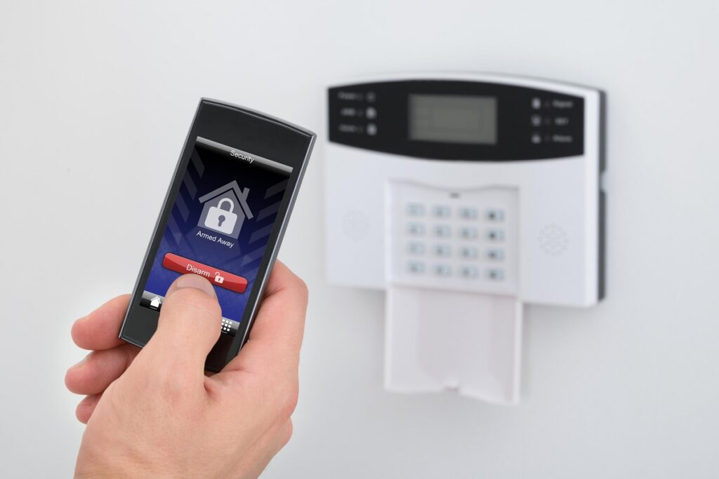 Una mano sostiene un smartphone con una aplicación de seguridad de alarma frente a un teclado de alarma doméstica instalado en una pared.