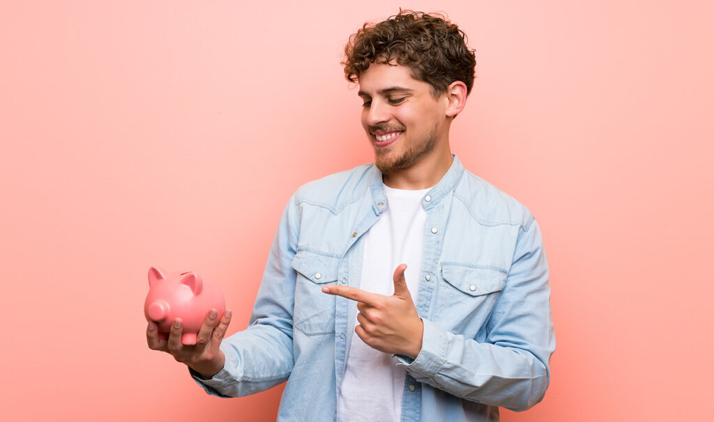 Hombre sonriente con chaqueta vaquera clara sosteniendo y señalando una hucha rosa sobre fondo rosa, promocionando cuentas de ahorro.