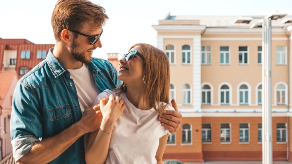 Una pareja sonriente con gafas de sol se abraza al aire libre delante de un edificio en un día soleado, disfrutando juntos del momento como una perfecta cuenta compartida.
