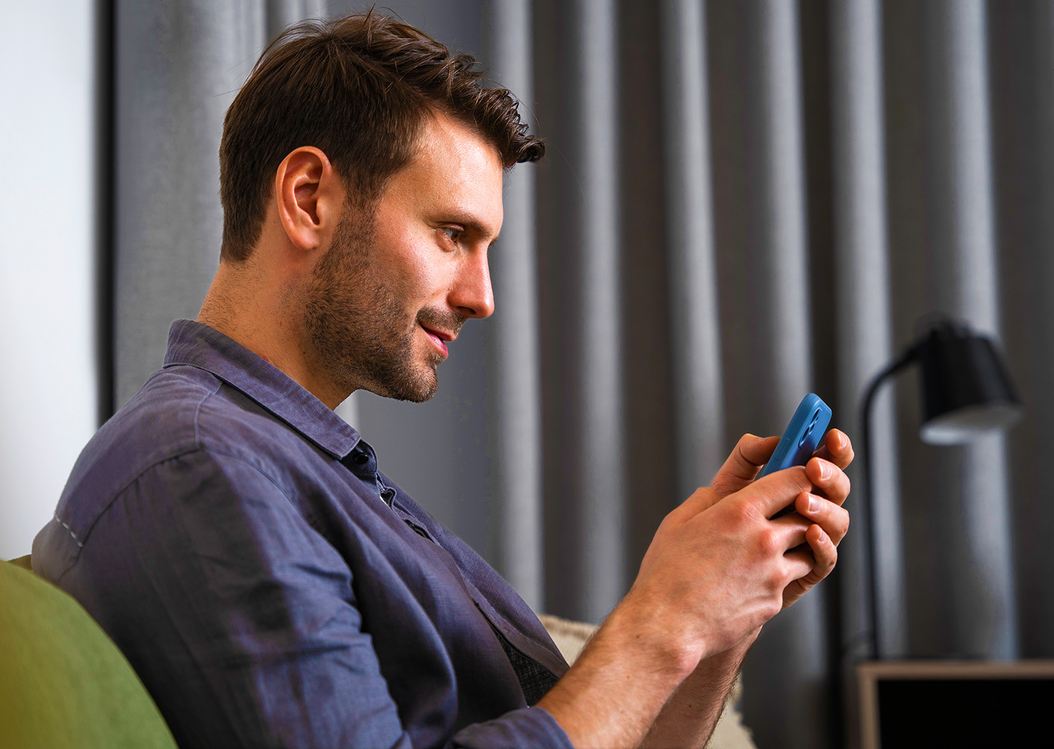 Hombre sentado en un sofá, sonriendo mientras utiliza su smartphone para explorar la nueva app de Banco Mediolanum, con cortinas grises de fondo.