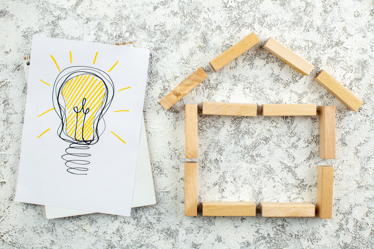 Dibujo de una bombilla junto al contorno de una casa sencilla hecha con bloques de madera sobre una superficie texturizada, que simboliza ideas como el acceso a la vivienda o las soluciones de expertos como José Elías.