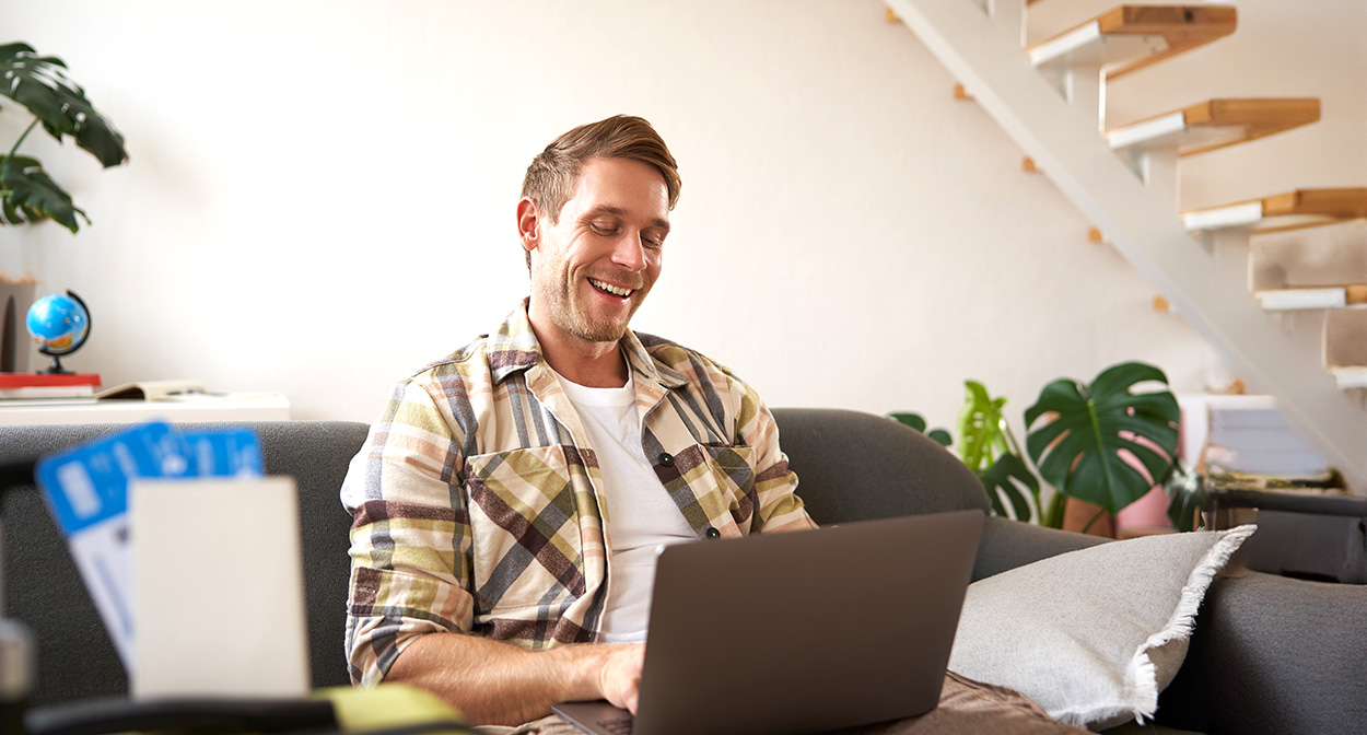 Hombre sonriente sentado en un sofá trabajando con un portátil en un luminoso y moderno salón, investigando la mejor cuenta para autónomos para su negocio por cuenta propia.