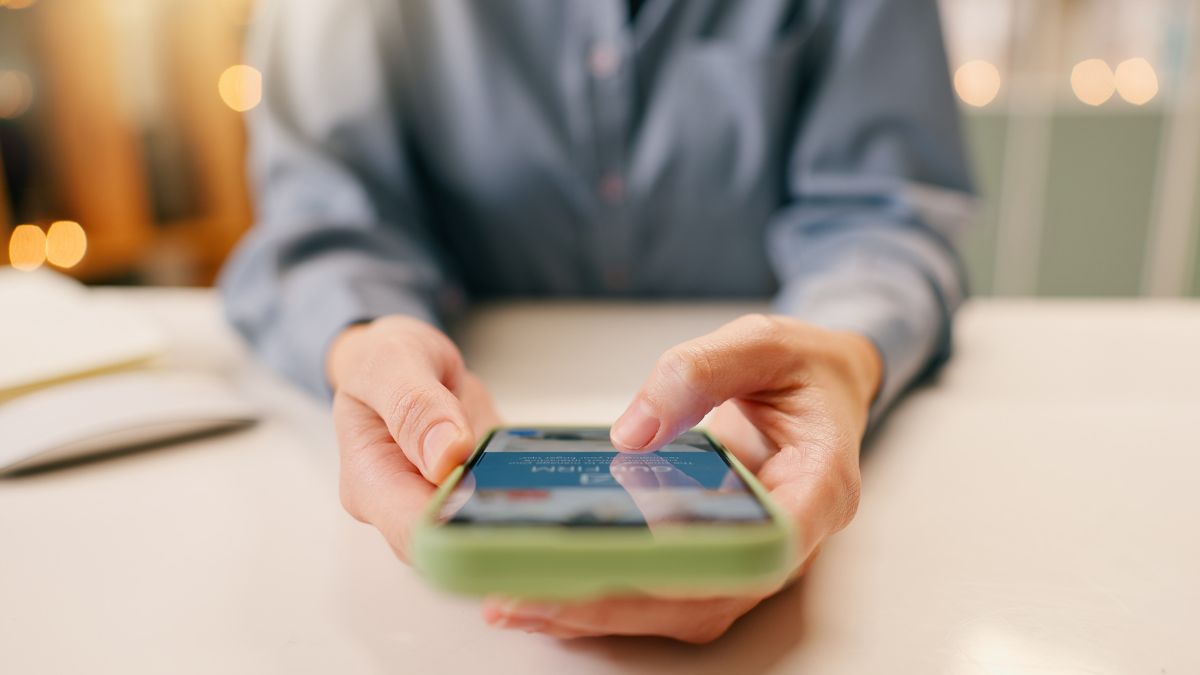 Primer plano de una persona utilizando un smartphone con ambas manos mientras está sentada en una mesa blanca, posiblemente revisando consejos financieros o aprendiendo los pasos para cobrar dinero de forma segura.