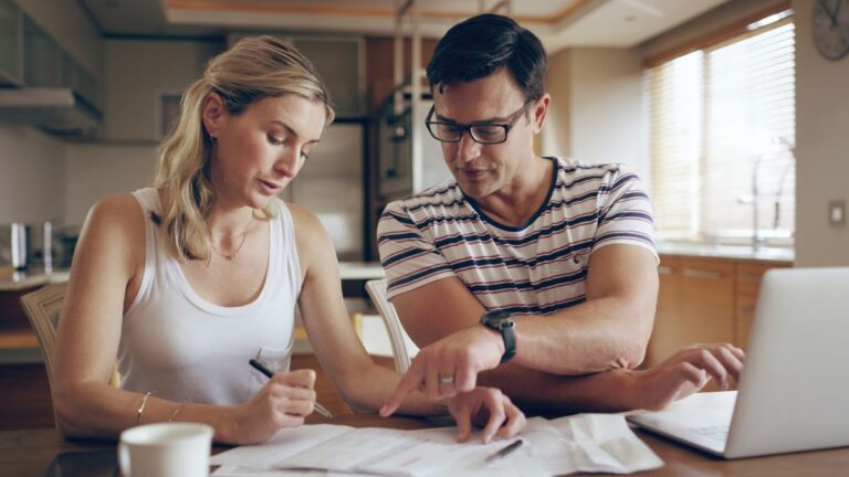 Una pareja se sienta a la mesa de una luminosa cocina, revisando papeles y discutiendo sobre la gestión del dinero con un portátil y una taza de café.