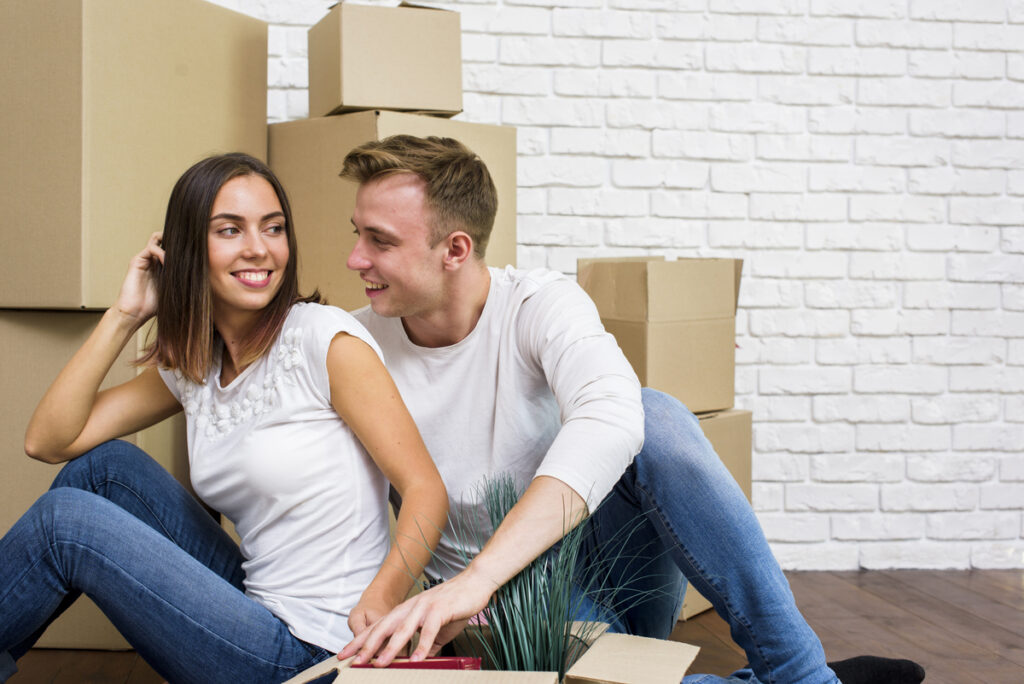 Una pareja sonriente se sienta en el suelo, rodeada de cajas de mudanza en su nuevo hogar, entusiasmada por empezar de cero tras conseguir hipotecas favorables.