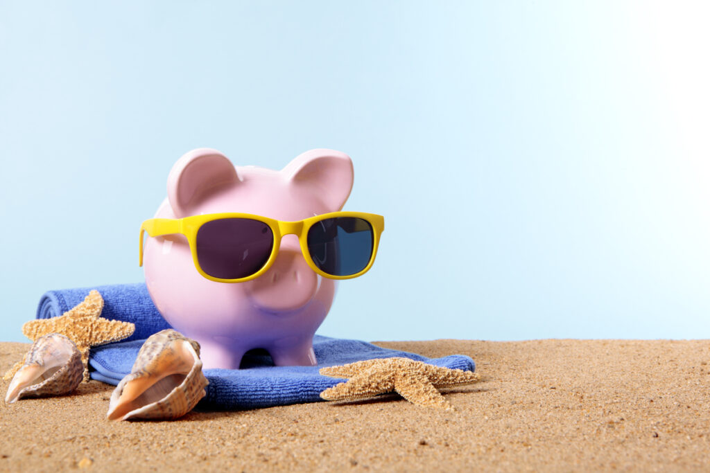 Hucha rosa con gafas de sol amarillas sobre arena con conchas, una toalla azul y un fondo azul claro, lista para las vacaciones.
