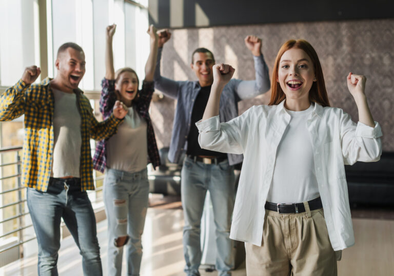 Cuatro adultos jóvenes entusiasmados celebran en el interior, levantando los brazos y sonriendo con entusiasmo, tal vez después de descubrir las mejores cuentas nómina para optimizar sus finanzas juntos.