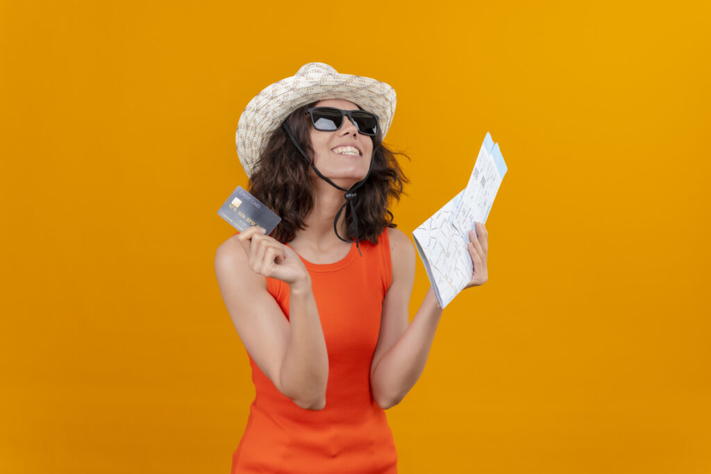 Mujer sonriente con sombrero y gafas de sol, sosteniendo una tarjeta de crédito y un mapa sobre un fondo naranja, lista para vivir aventuras sin tener que pagar un suplemento.