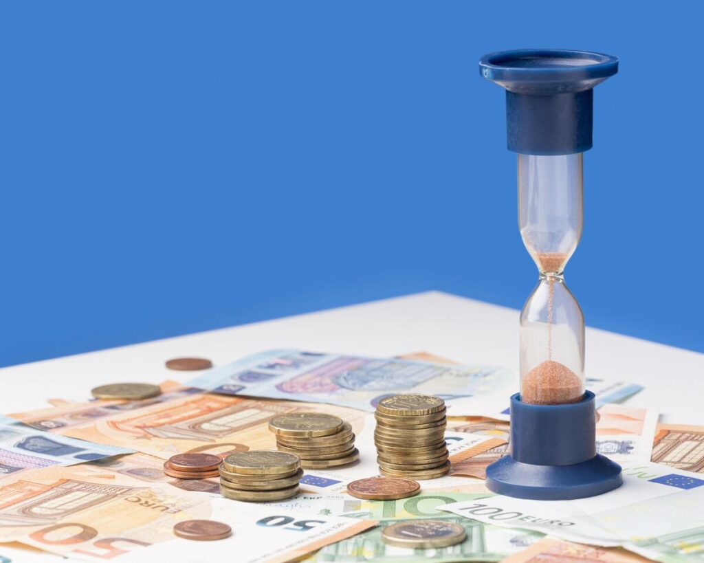 Un reloj de arena con arena, billetes de euro y monedas sobre una mesa, que simbolizan el tiempo y el dinero: un recordatorio de cómo los depósitos al 3 % pueden ayudarte a hacer crecer tus ahorros de forma eficaz.