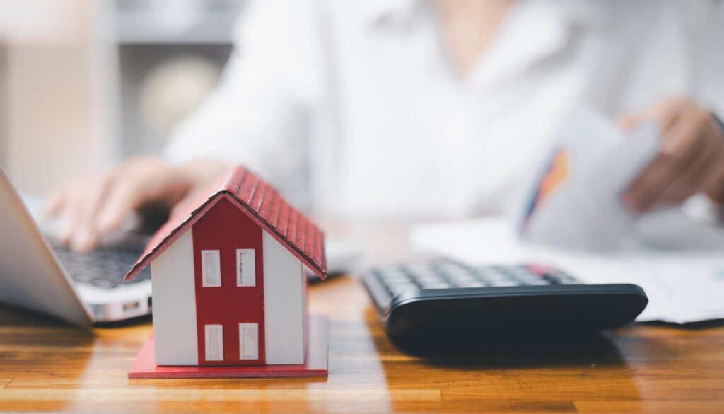 Una casita, una calculadora y una persona con un ordenador portátil y unos papeles, lo que sugiere temas relacionados con las finanzas o la planificación hipotecaria, como «cuándo cambiar de hipoteca» o «ahorrar con la hipoteca».
