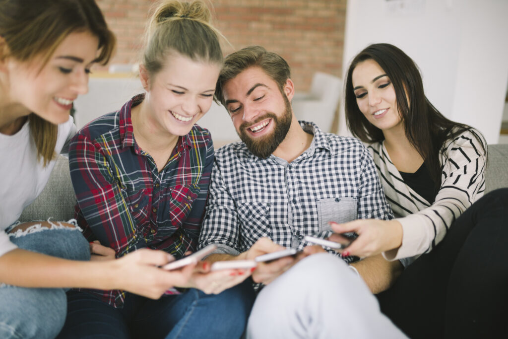 Cuatro amigos sentados en un sofá, riendo y mirando juntos sus móviles, mientras hablan de finanzas y comparten consejos sobre cómo algunos inversores están ahorrando 61 euros al mes.