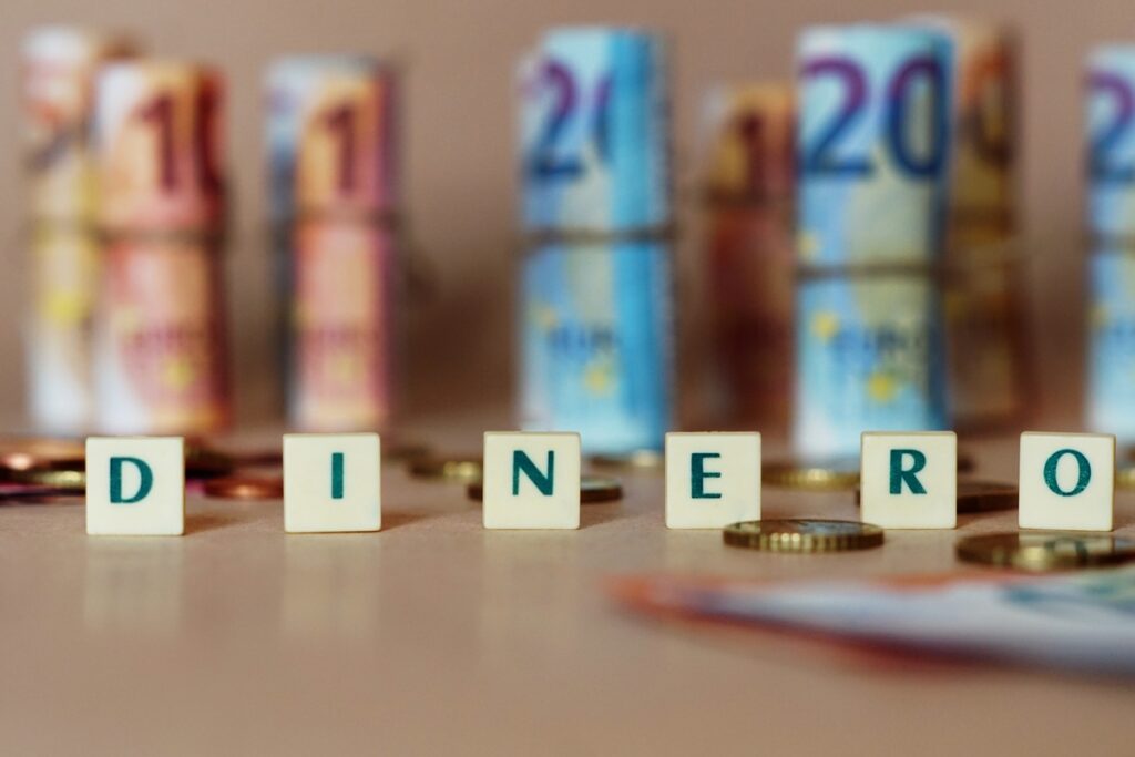 Las fichas de Scrabble forman la palabra «DINERO», con billetes y monedas de euro difuminados, lo que evoca los ahorros de una cuenta nómina.